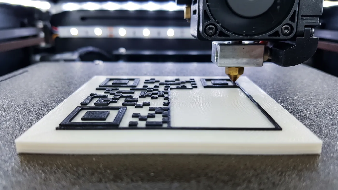 Boquilla de una impresora 3D depositando filamento negro sobre una placa blanca ya impresa, formando un código QR en relieve